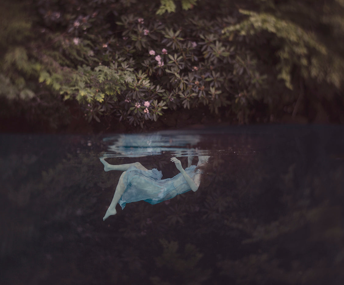 An ethereal photograph of a person lying on their back in a body of water with flowers and foliage around, representing a whimsical and mystical artistic style.