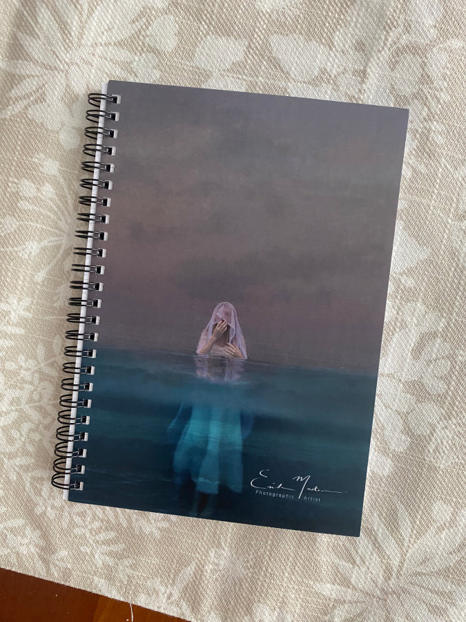 A spiral-bound notebook with a custom cover featuring a fine art photograph of a girl standing in the ocean during sunset, with the name 'Erika masterson' inscribed on the artwork.