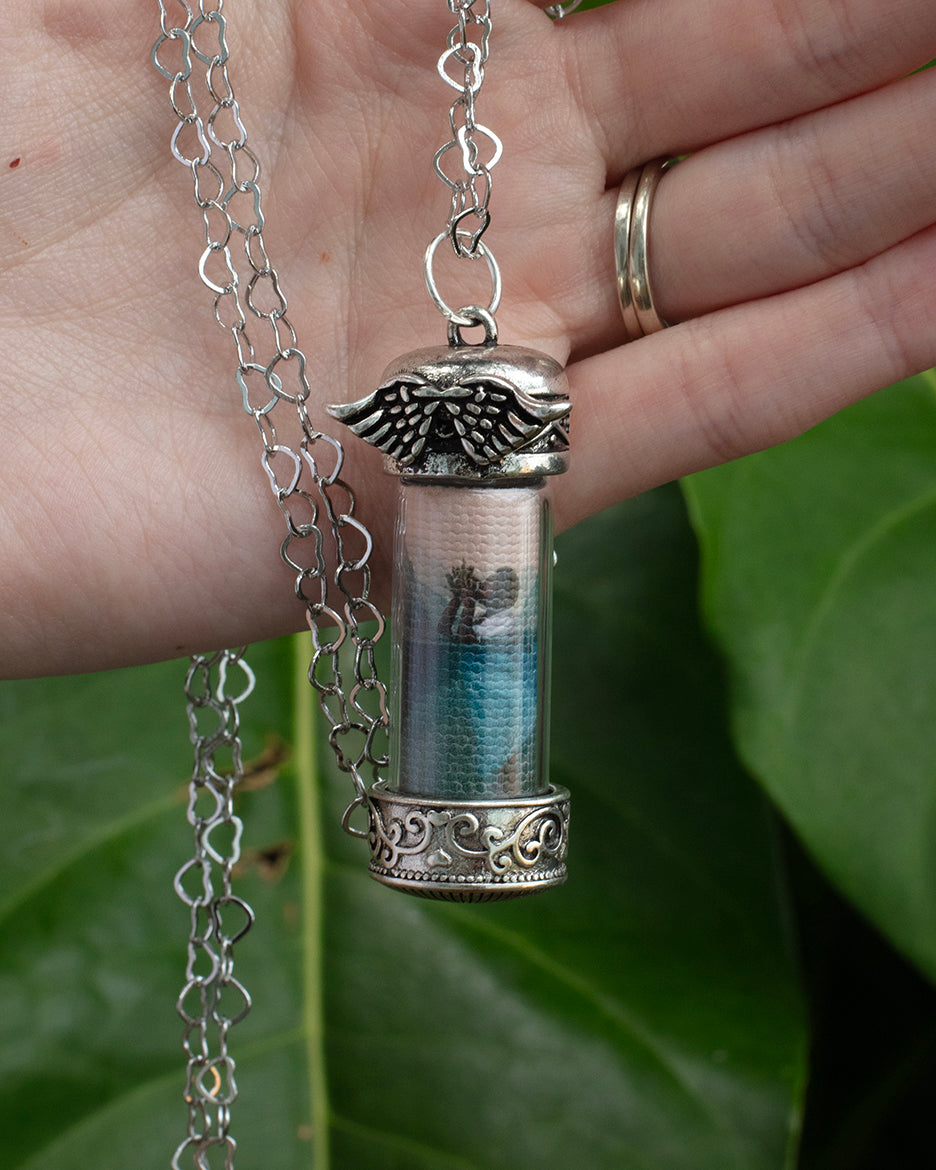 A glass capsule photo necklace with a silver metal chain and adjustable length. The capsule contains a photograph of a woman with a sextant in a mountain lake setting.