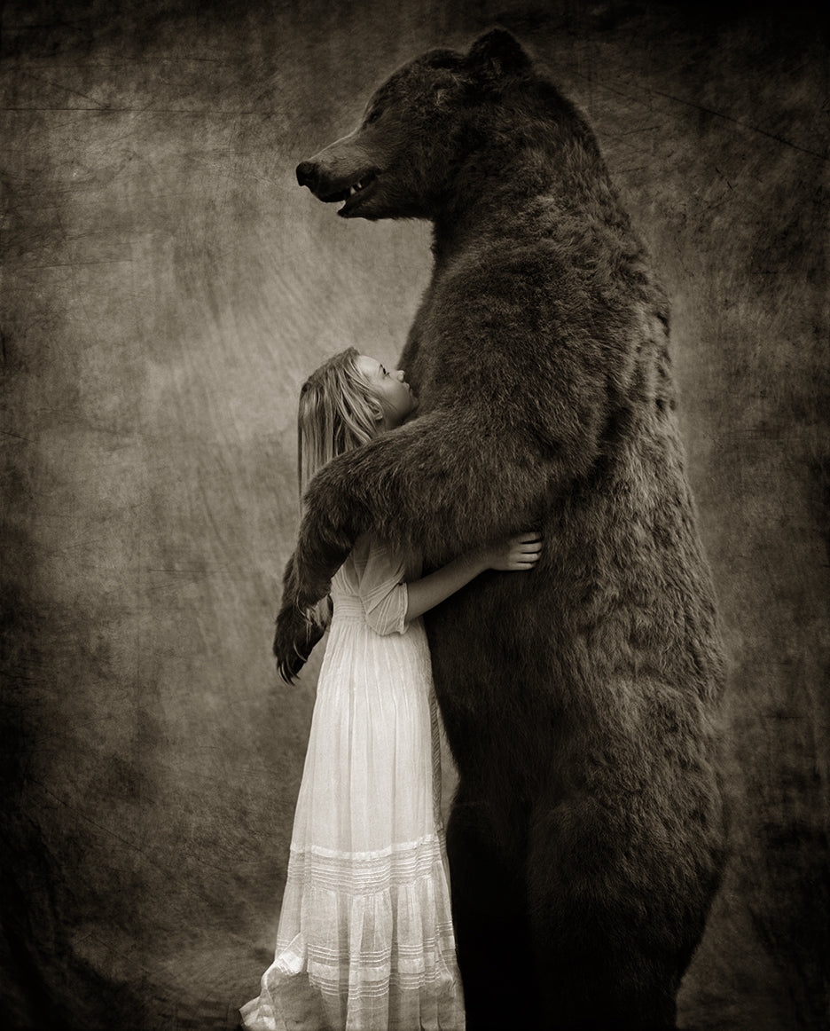 A black and white photograph of a girl cradled in the arms of a bear, with the girl looking up at it.