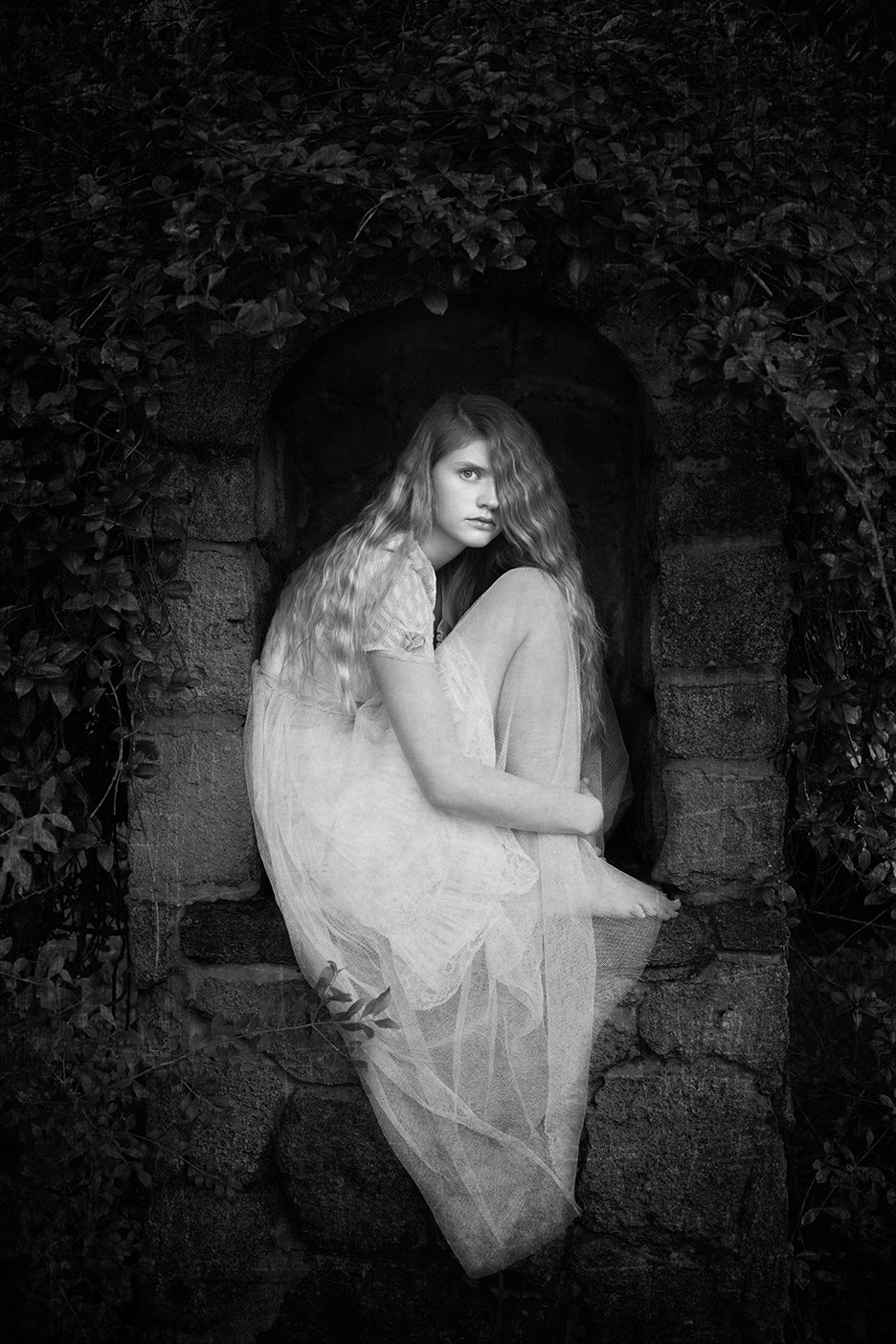 A black and white fine art photograph featuring a woman with mysterious eyes, cradled in the embrace of a small cave, surrounded by darkness and with a hint of light coming through the trees outside.