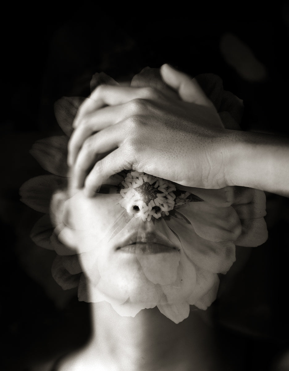 A close-up black and white photograph of a woman's face with a delicate flower superimposed on it, covering the lower part of the face.