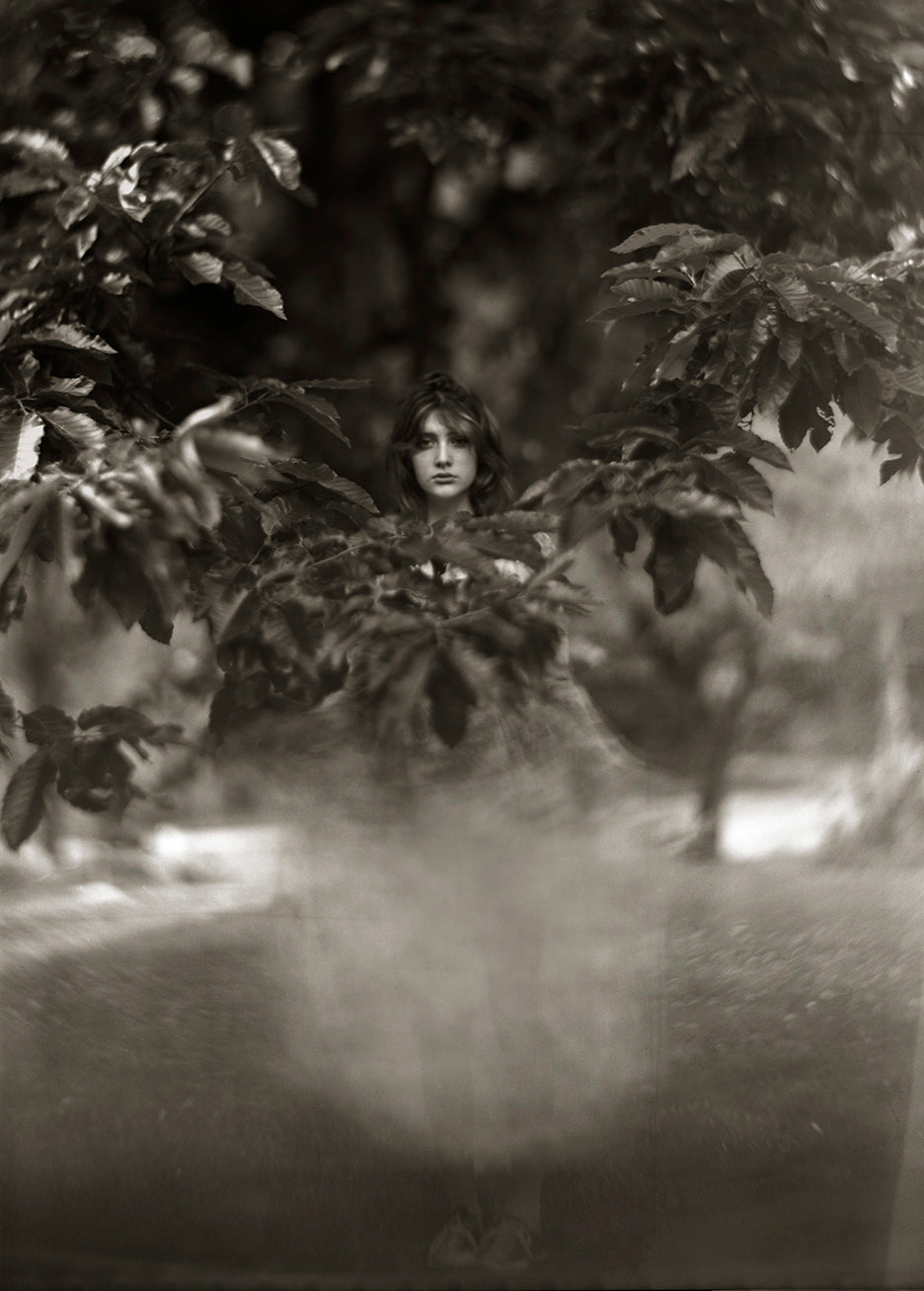 A black and white photograph of a girl standing beneath the branches of a chestnut tree, with a blurred background.