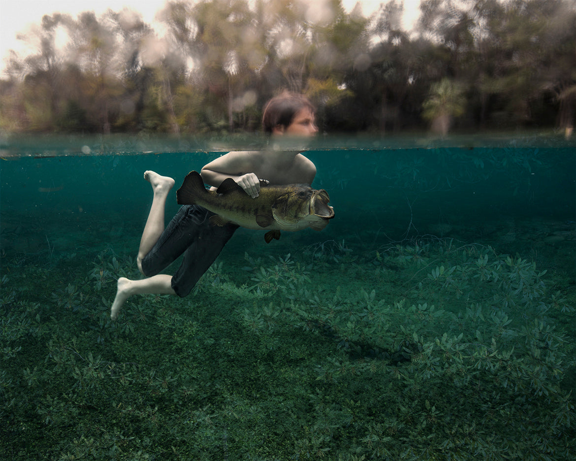 A photograph featuring a young boy swimming in clear water, holding onto a large bass fish, with a blurred natural background.
