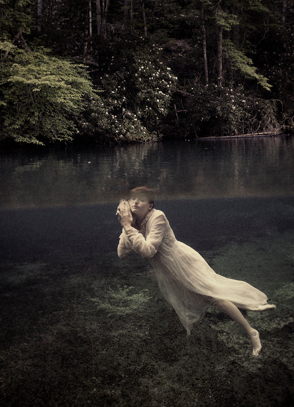 A photograph of a woman underwater, cradling a conch shell to her ear, with a blurred natural background.