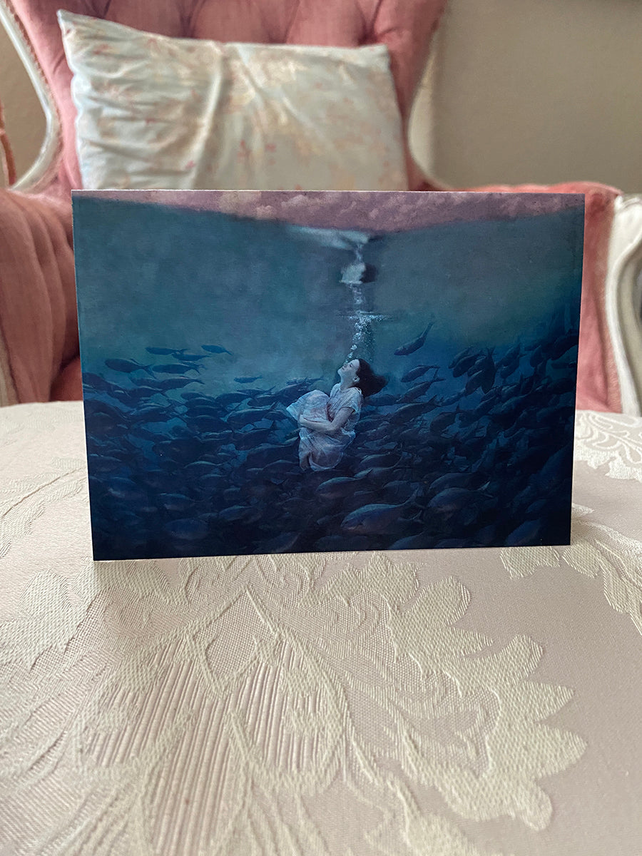 A note card with a fine art photograph of a girl underwater, surrounded by playful fish and bubbles. The card is presented on a table with a cushion in the background.