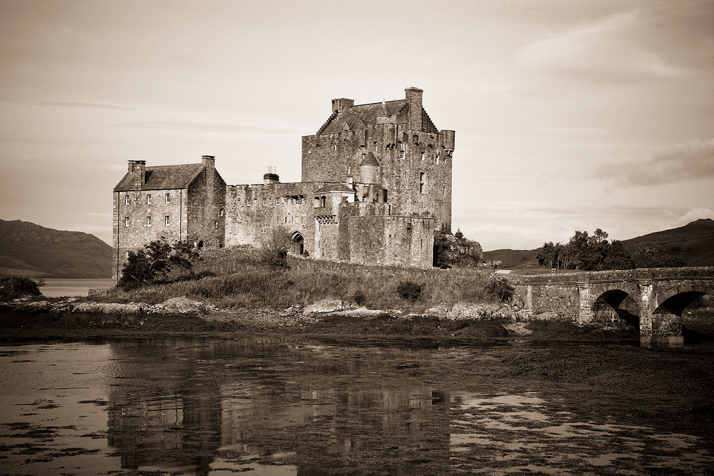 A black and white photograph of Eilean Donan Castle, a Scottish castle situated on an island with a bridge leading to it, surrounded by water and a cloudy sky in the background.