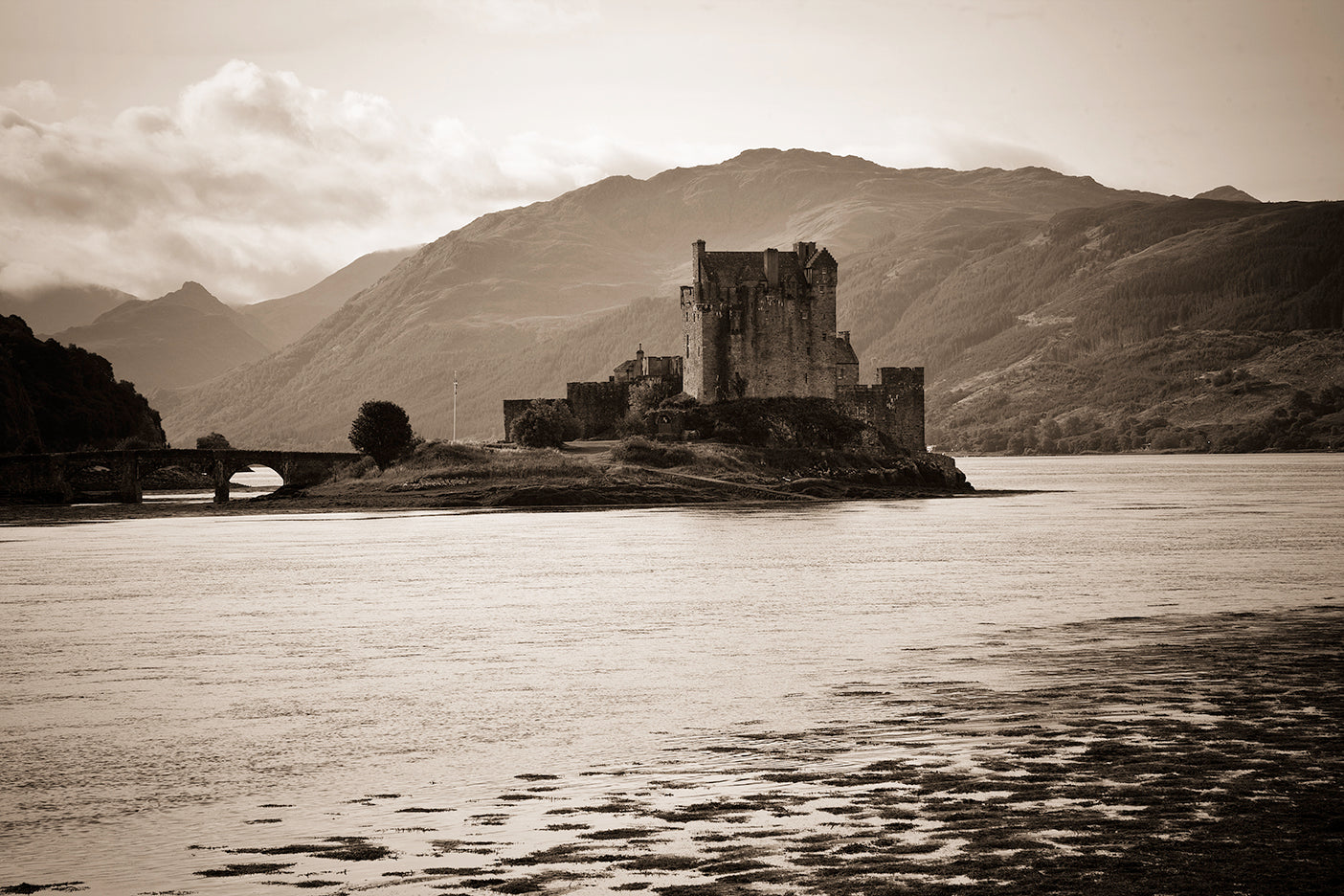 Black and white photograph of Eilean Donan Castle located on a Loch with surrounding mountains in the background.