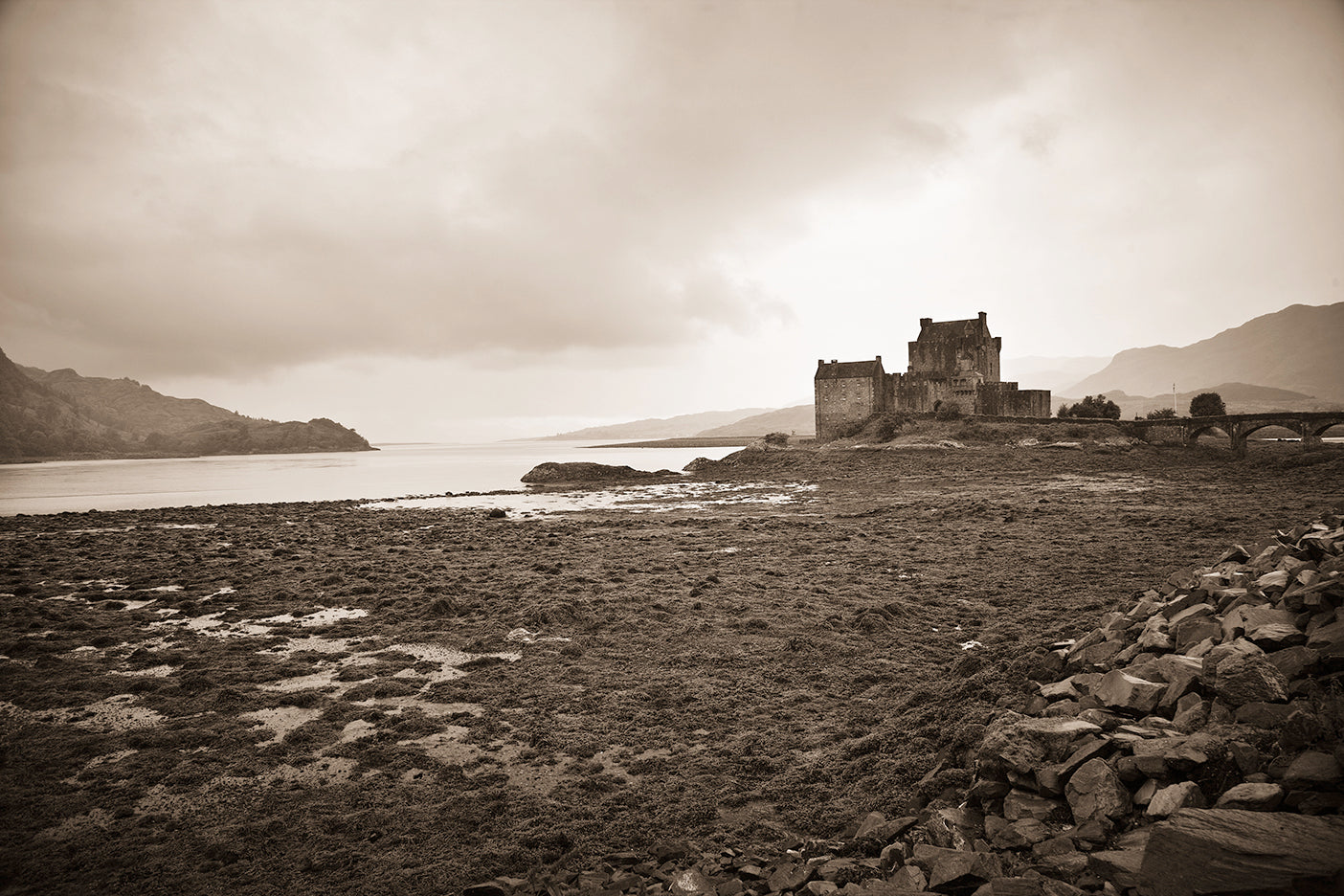 A black and white photograph of Eilean Donan Castle, located on a Loch with a cloudy sky above and rugged terrain in the foreground.