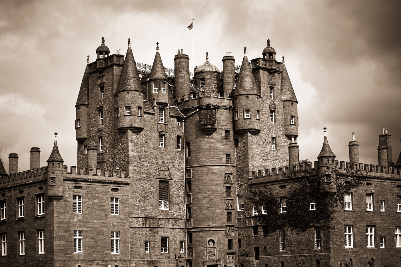 A black and white image of Glamis Castle, showing the front facade with multiple towers and spires against a cloudy sky.