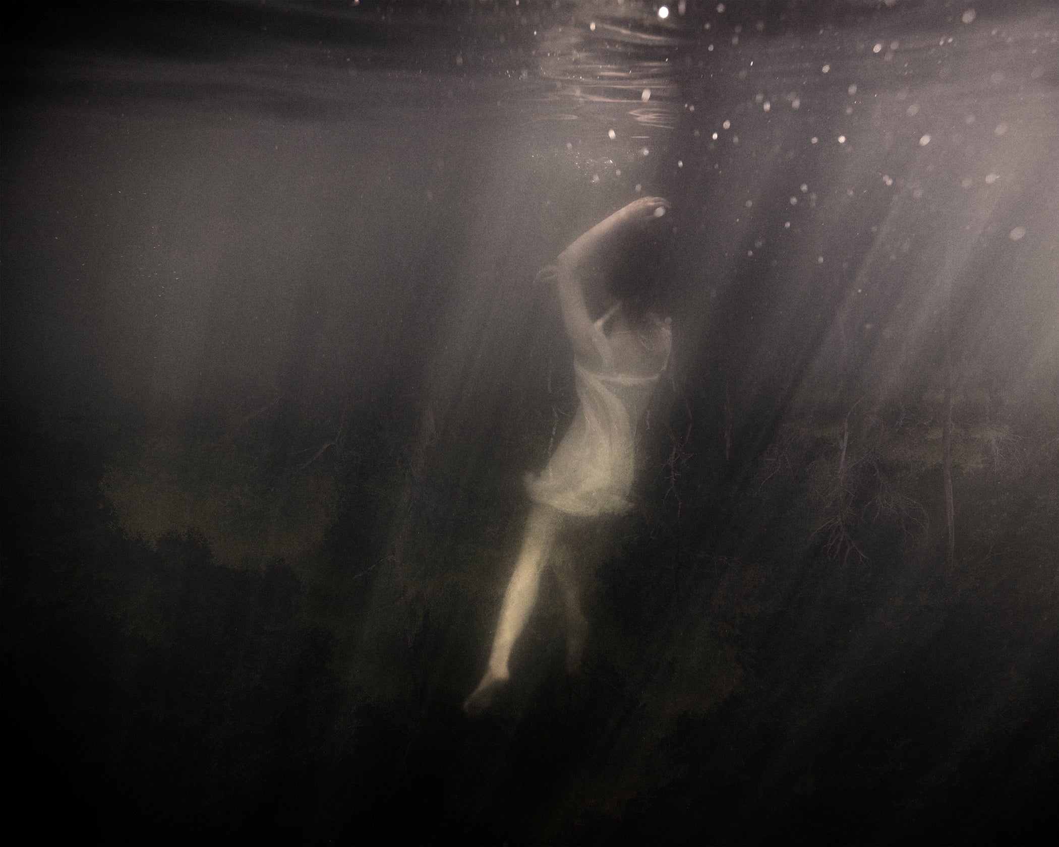 An underwater photograph of a person in a dress reaching upwards, with light beams and a generally serene underwater scene.