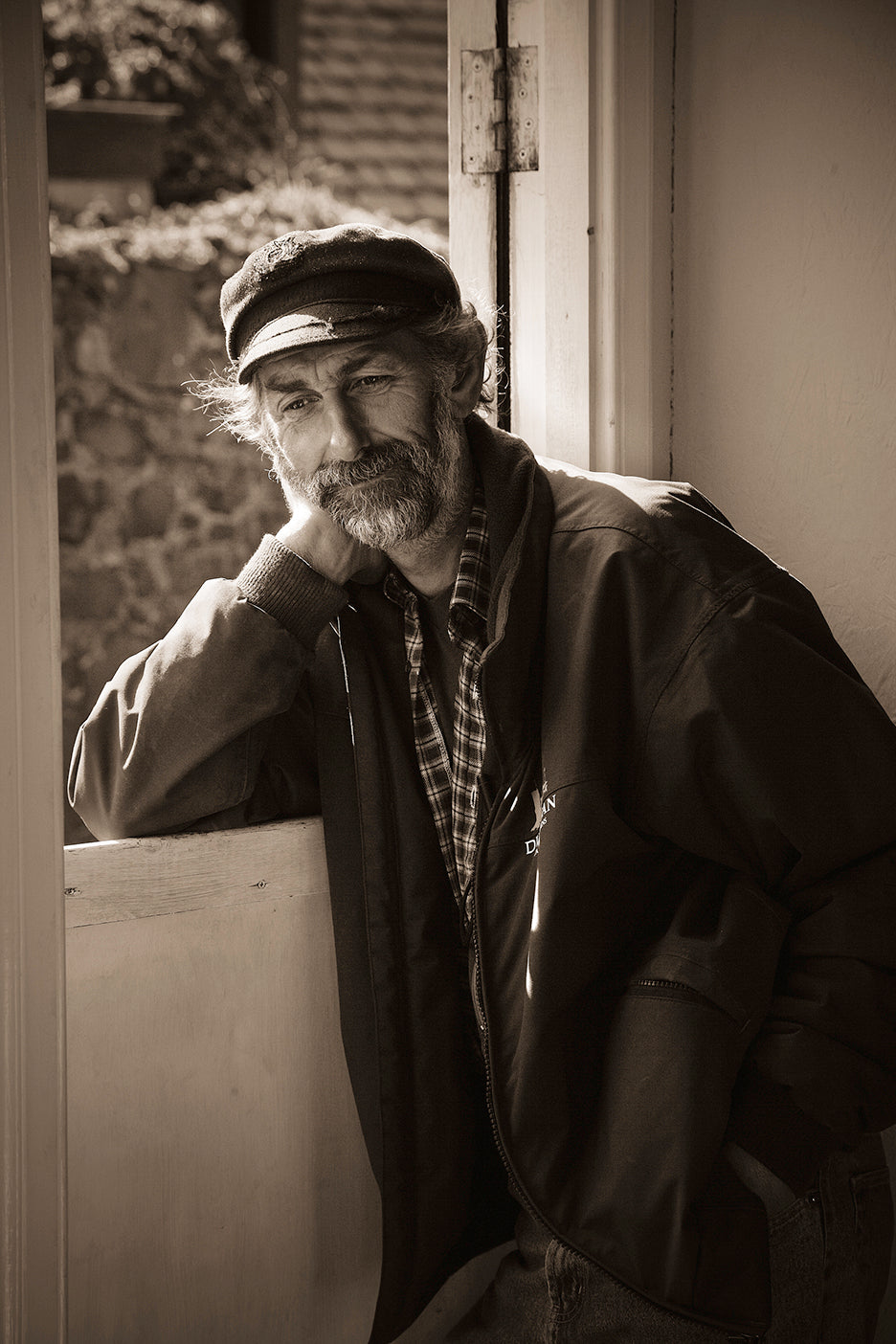 Black and white photograph of a Scottish fisherman sitting by a door, wearing a hat and a jacket, with a wall adorned with framed photographs in the background.
