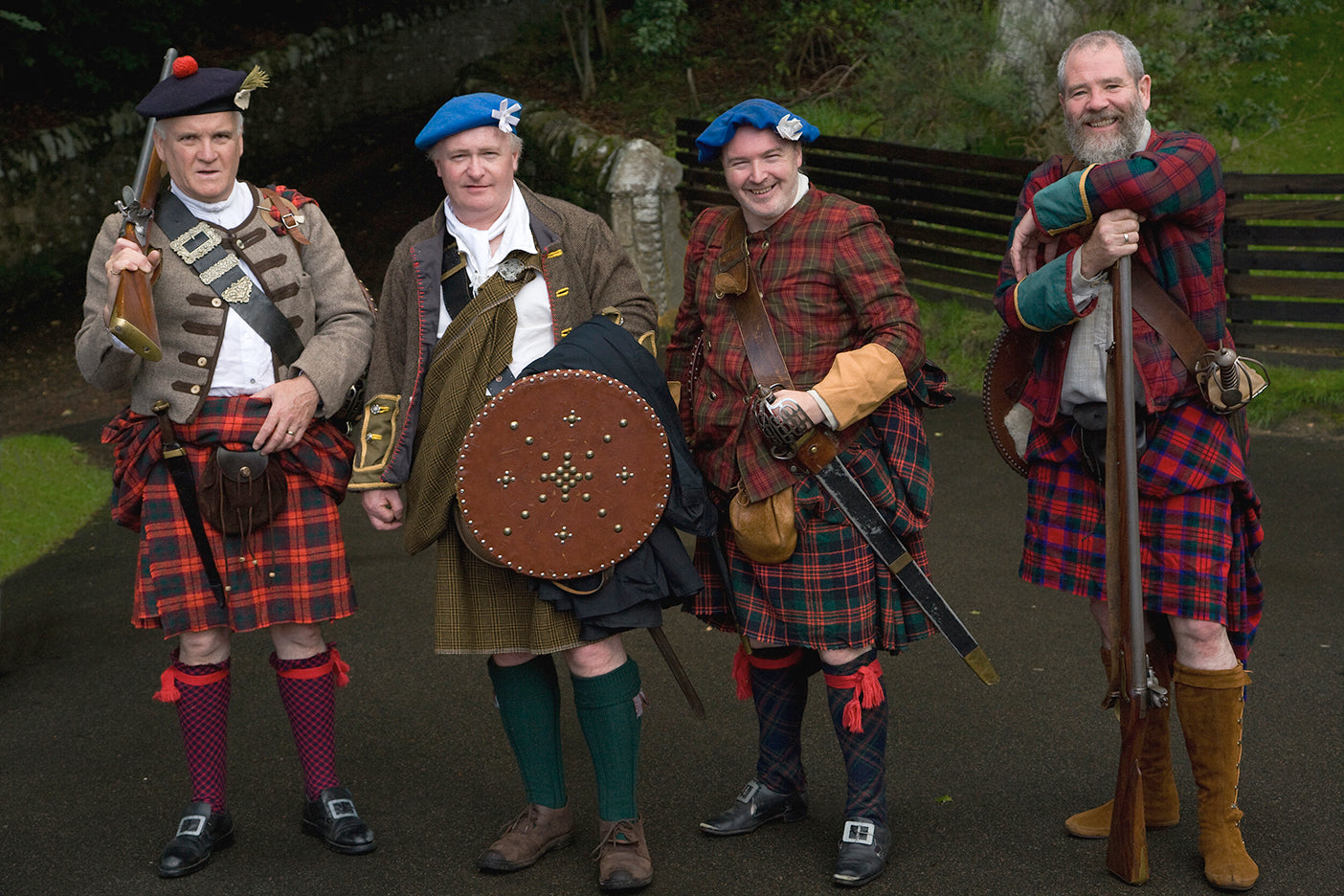 Four men dressed in Scottish kilts and traditional attire, standing in front of a castle.