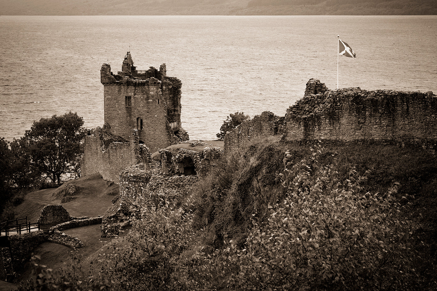 Black and white photograph of Urquhart Castle with a flag flying in the distance, set against a body of water.