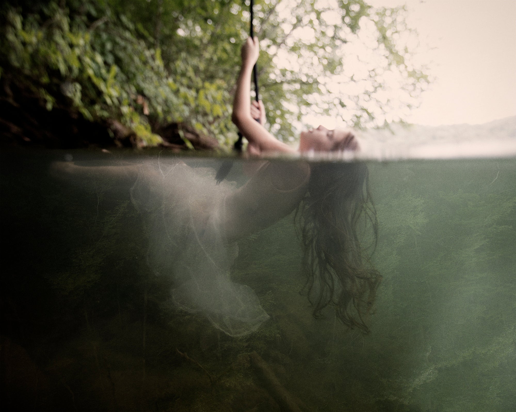 An artistic representation of a person floating underwater with a rope attached to them, with above-water foliage in the background.