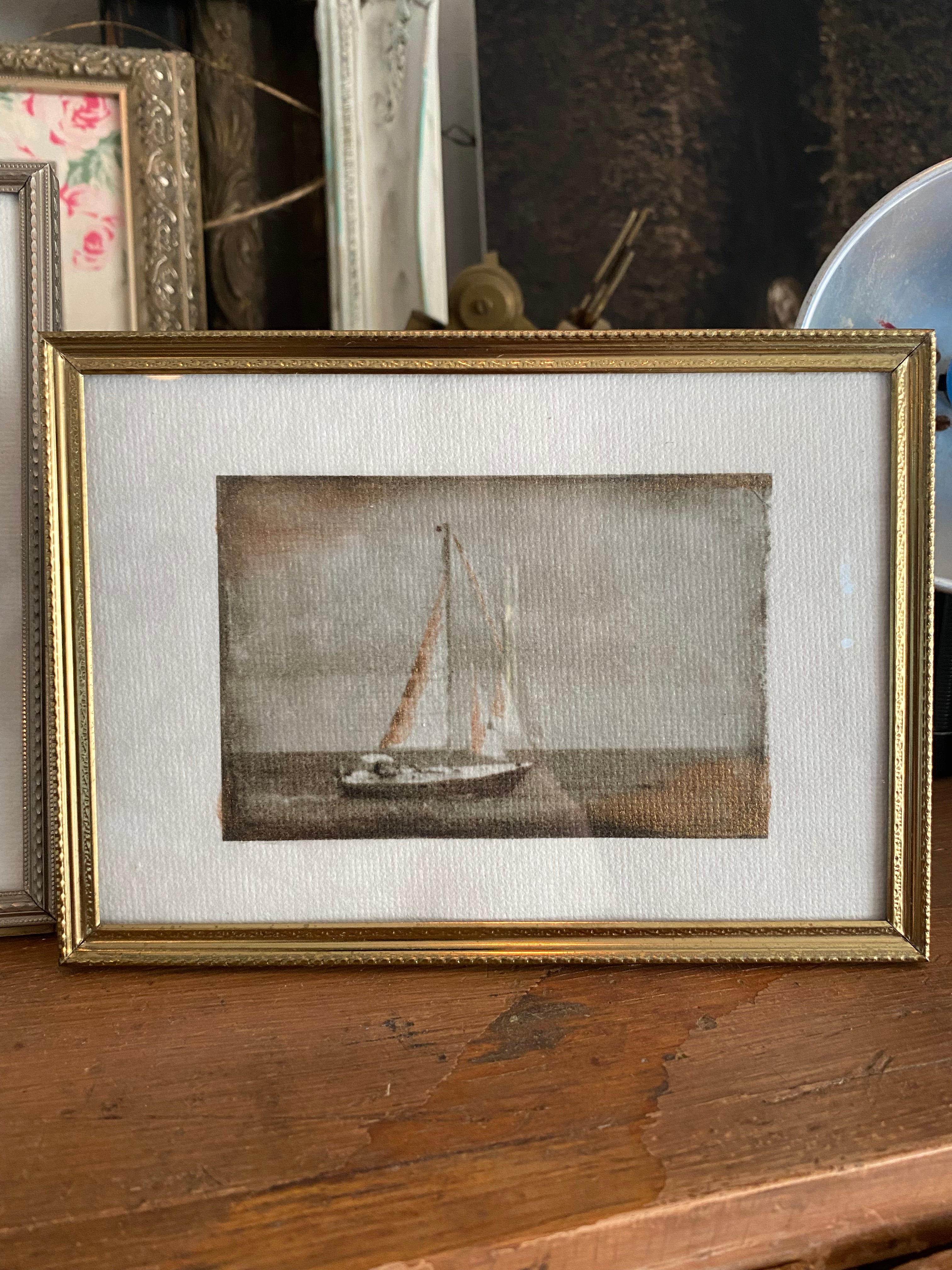 A mini hand-painted artwork depicting a sailboat, framed in a vintage gold frame, set against a backdrop of various framed items on a wooden surface.