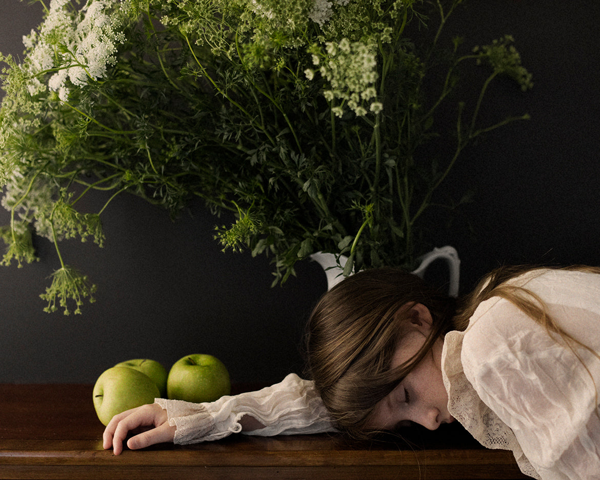 A piece of fine art photography featuring a person with their head resting on a surface, surrounded by Queen Annes lace and apples, evoking a sense of peace and connection with nature.