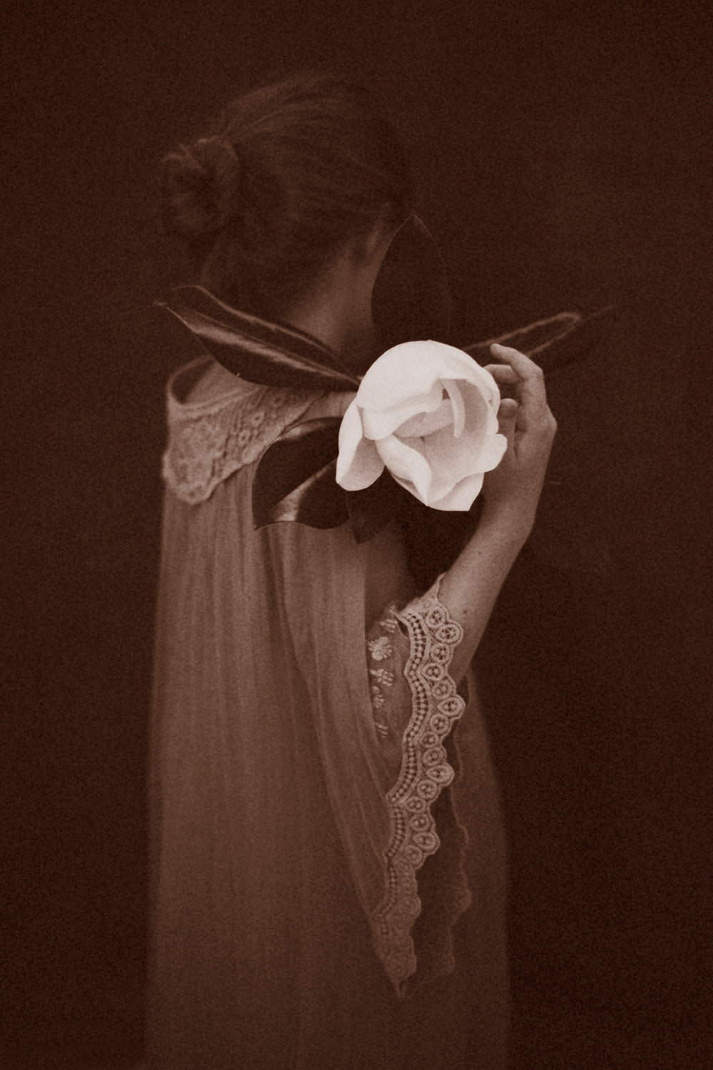 A black and white photograph of a person from behind, holding a magnolia flower close to their face, wearing a dress with lace details.