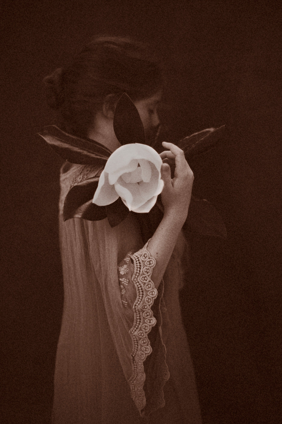 A black and white photograph of a person holding a magnolia flower close to their face, with a soft focus on the flower and a blurred background.
