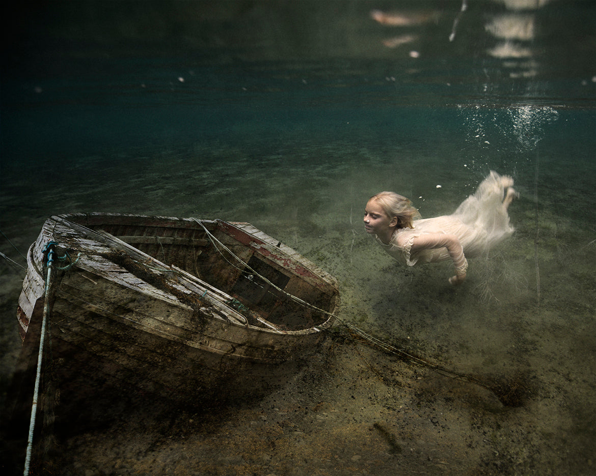 A fine art photograph featuring a young girl swimming underwater towards a boat on the ocean floor.