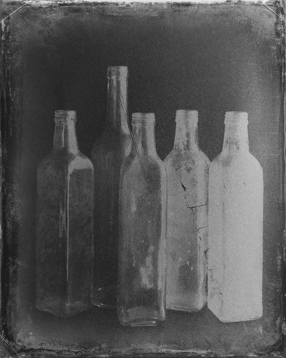 A black-and-white fine art photograph featuring a group of bottles, each with a unique shape, arranged in a way that creates a visual dialogue. The image emphasizes the contours and textures of the bottles, with an interplay of light and shadow.