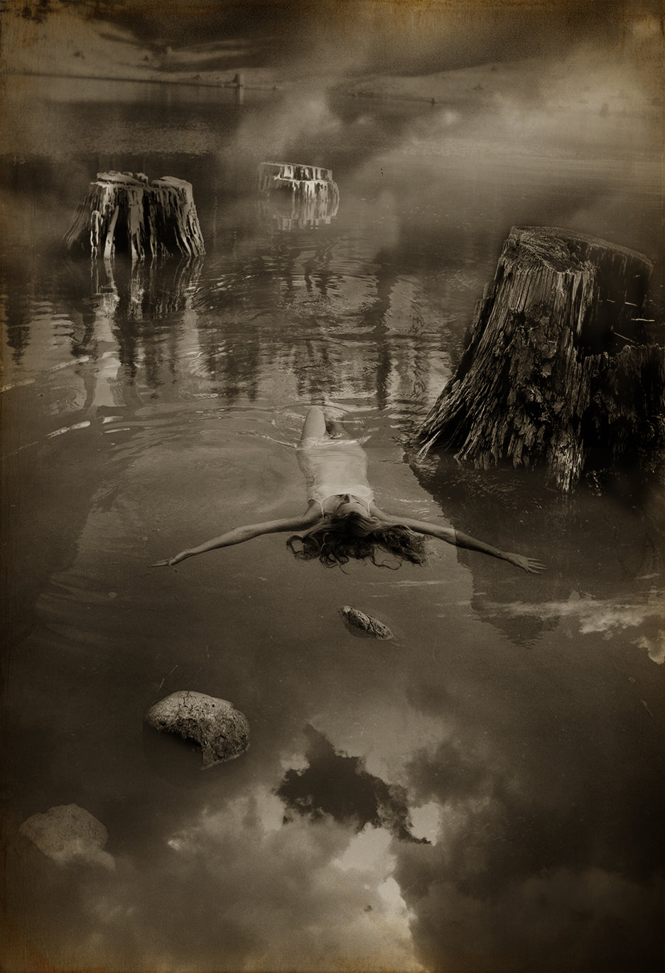 A black and white photograph of a person floating on water, with natural elements like trees and rocks in the background, creating a serene and dreamlike atmosphere.