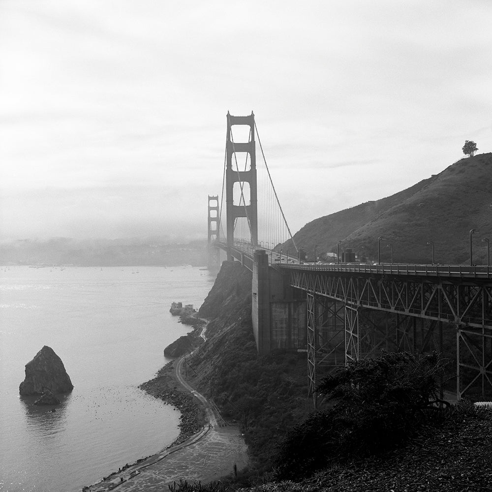 Black and white photograph of the Golden Gate Bridge with a misty background and a rock formation in the foreground on the left side.