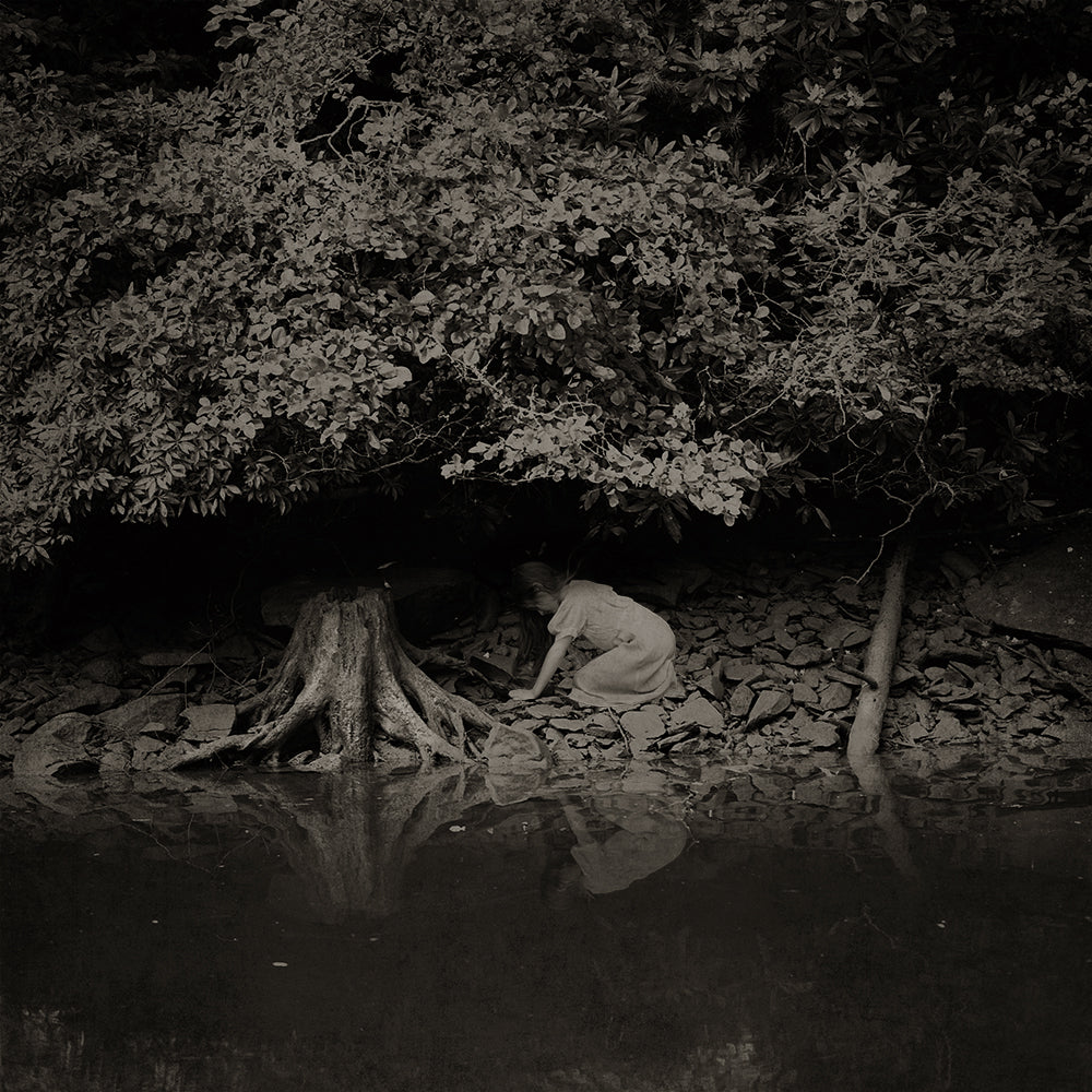 A black and white photo featuring a person crouched down by a body of water, with trees and a reflective surface in the background.