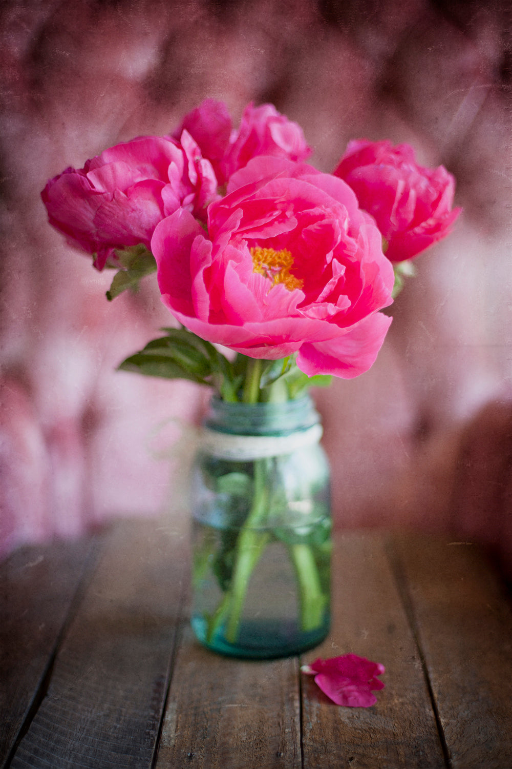 A photograph of a vase containing pink peonies, placed on a wooden surface with a blurred pink background.