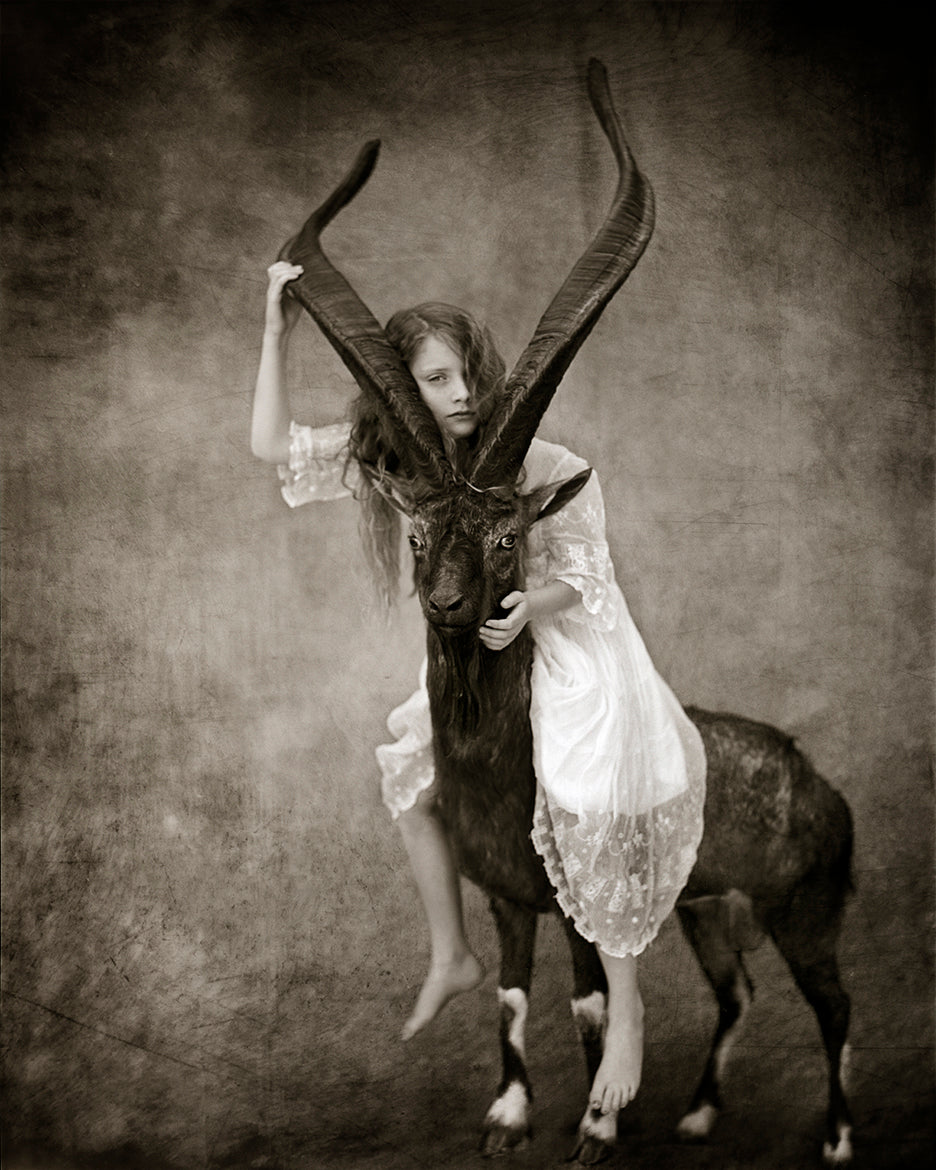 A black and white photograph featuring a young girl sitting atop an apex mountain goat, with a sepia-toned finish.