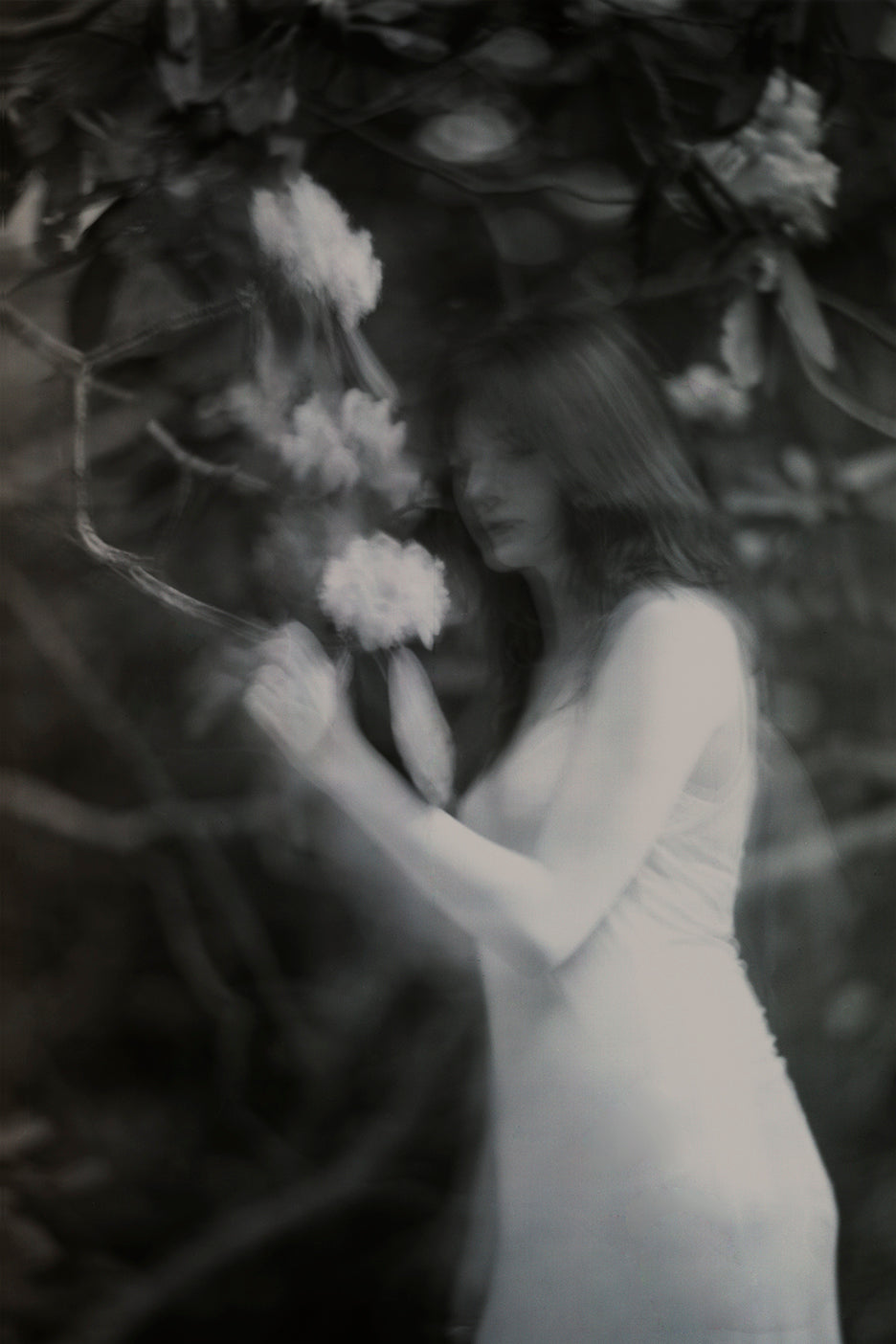 A black and white photograph featuring a young woman among trees with an ethereal and dreamlike quality.