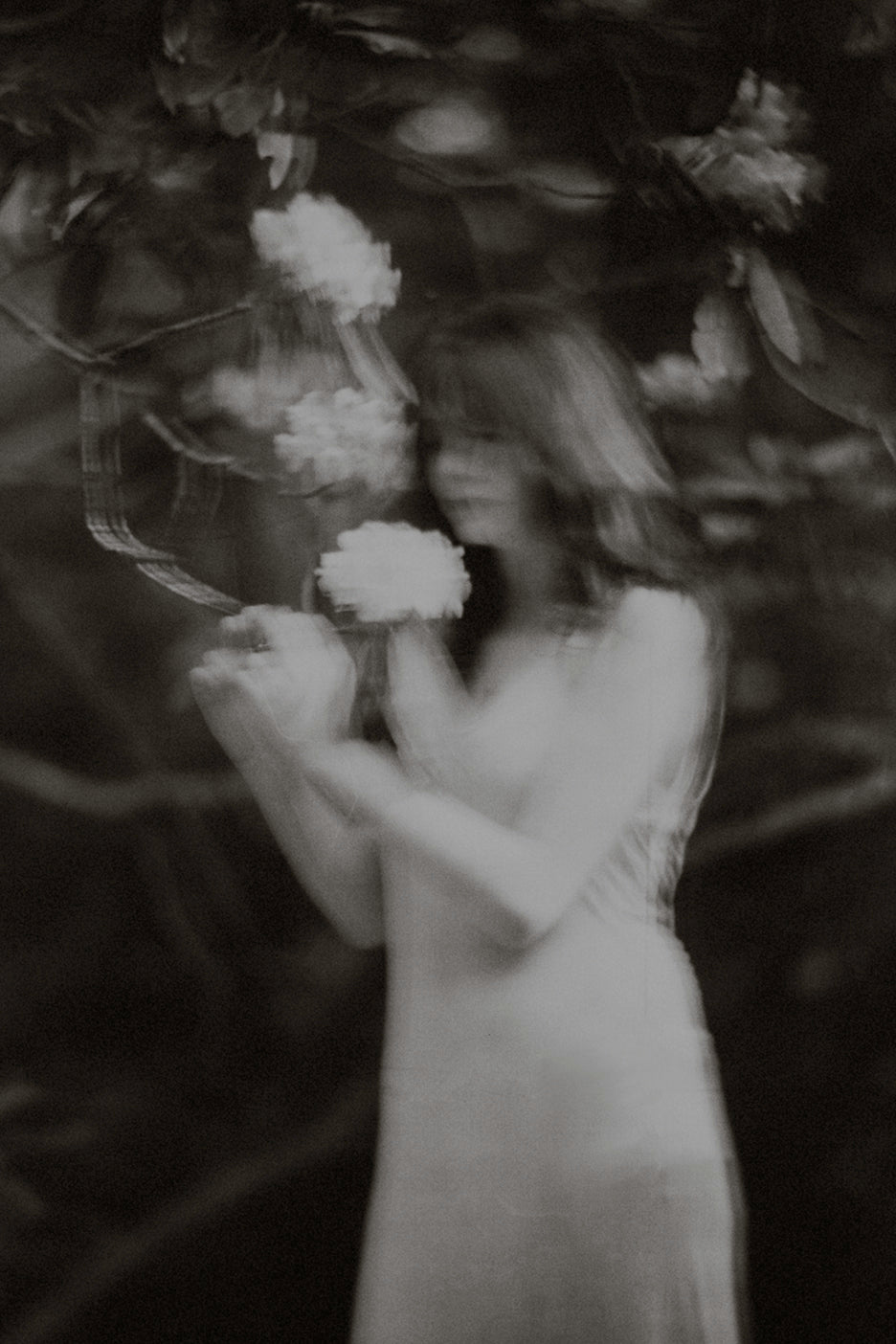 A black and white photograph of a young woman among trees and foliage, with a dreamlike and ethereal quality.