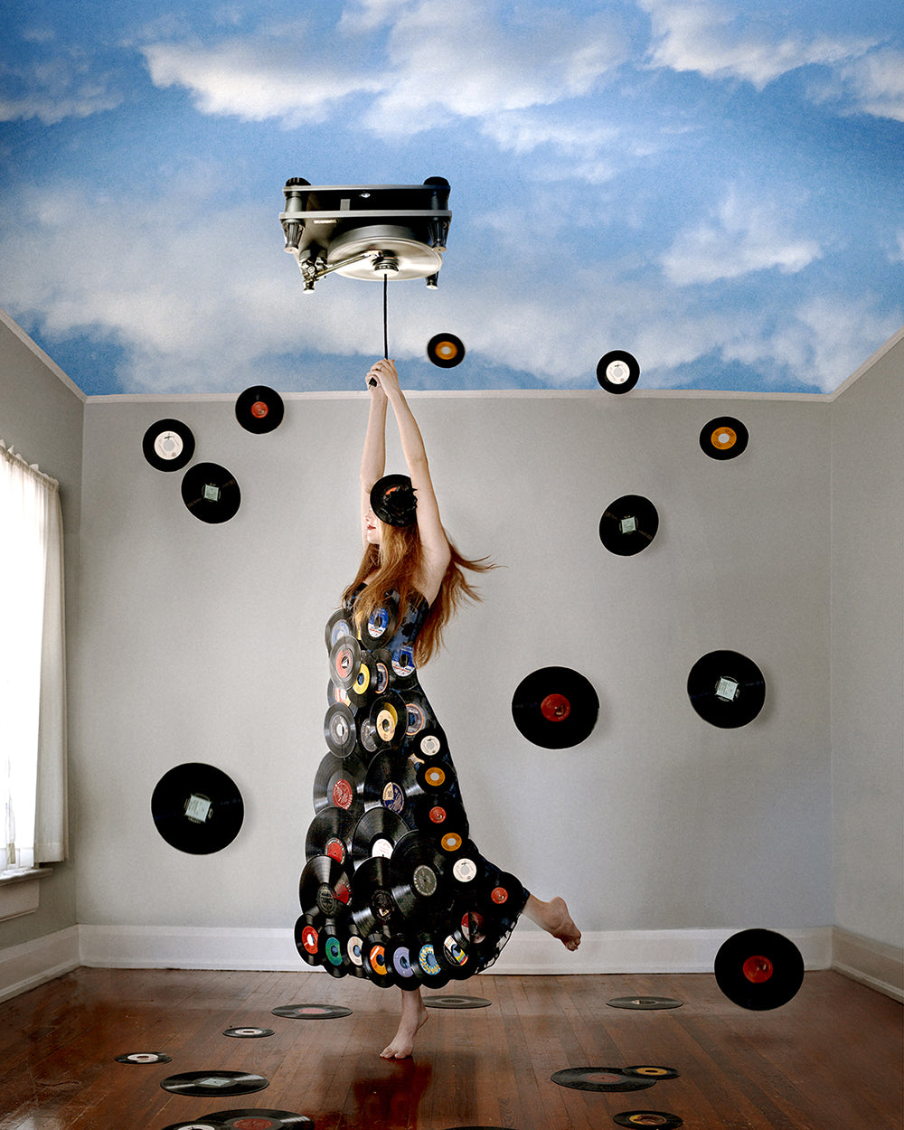 A woman in a dress with record patterns is suspended in the air in a room with a turntable above her and walls adorned with floating records. The room has a blue painted ceiling with clouds.