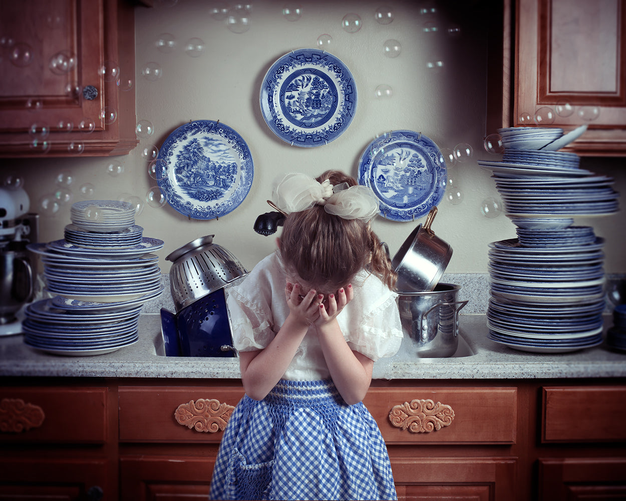 A young child with their hands covering their face, standing in a kitchen setting surrounded by stacked plates and cups, with blue and white decorative patterns.