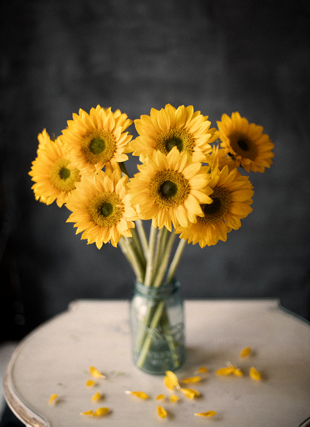 A bouquet of yellow sunflowers in a clear glass vase, with the flowers slightly tilted to show the back of the heads, set against a dark background.
