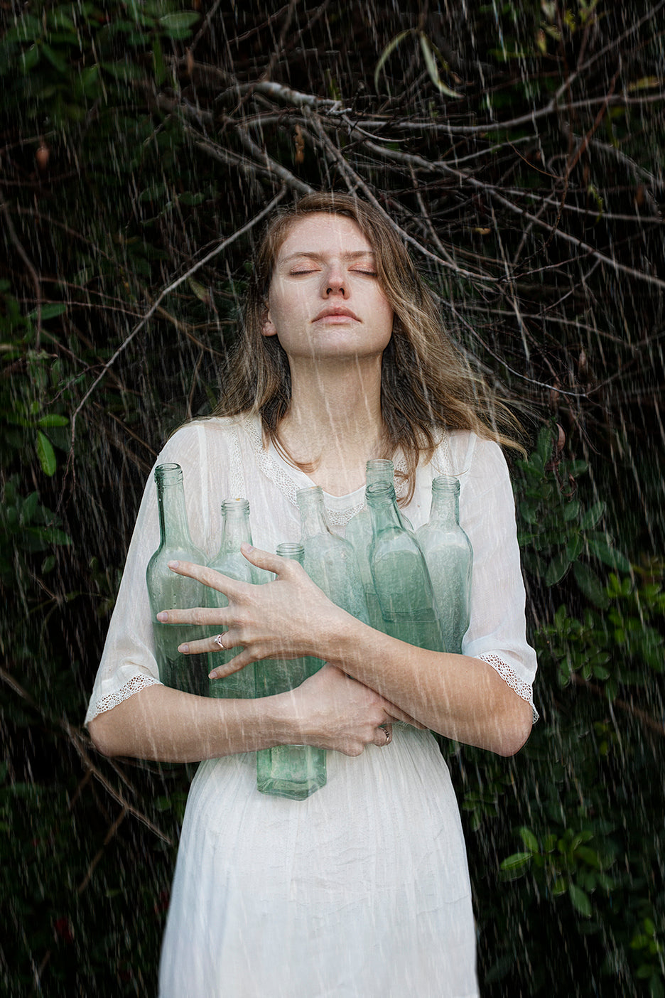 A person in a white dress holding green glass bottles, with a backdrop of nature.