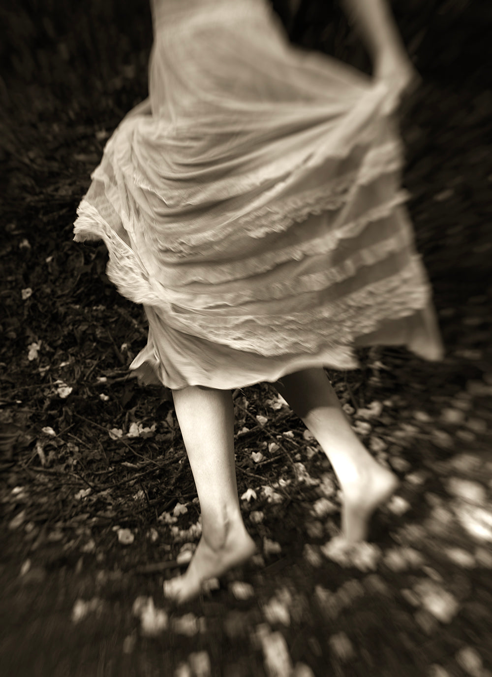 Black and white photograph of a person's lower legs and feet moving in a dancing pose with one foot lifted off the ground, surrounded by blurred foliage.