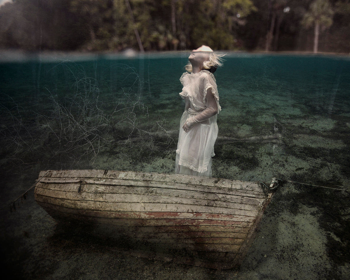 A black and white fine art photograph featuring a girl underwater with a sunken boat above her, rising toward the surface.