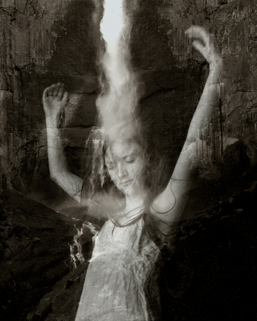 A black and white photo of a person with their arms raised, standing in front of a waterfall.
