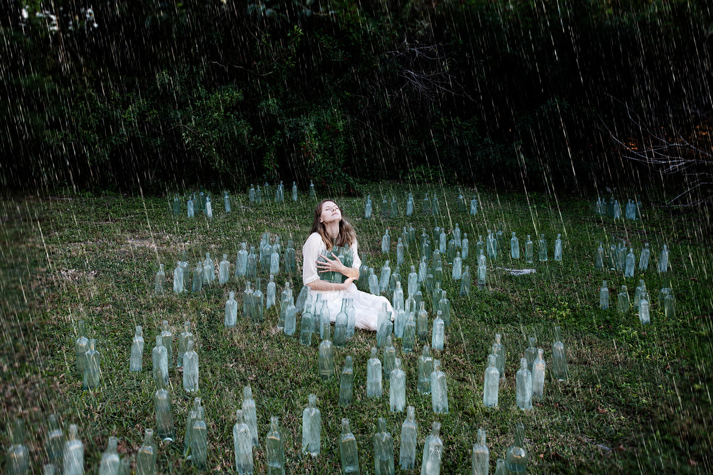Woman sitting among rows of glass bottles in a field during rain.